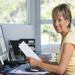 Woman in home office with computer and paperwork smiling Woman in home office with computer and paperwork smiling